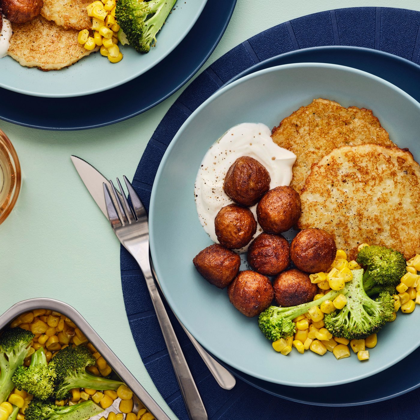 Su un tavolo, due ciotole blu contenenti polpette vegetali HUVUDROLL, frittelle di patate e un mix di mais e broccoli.