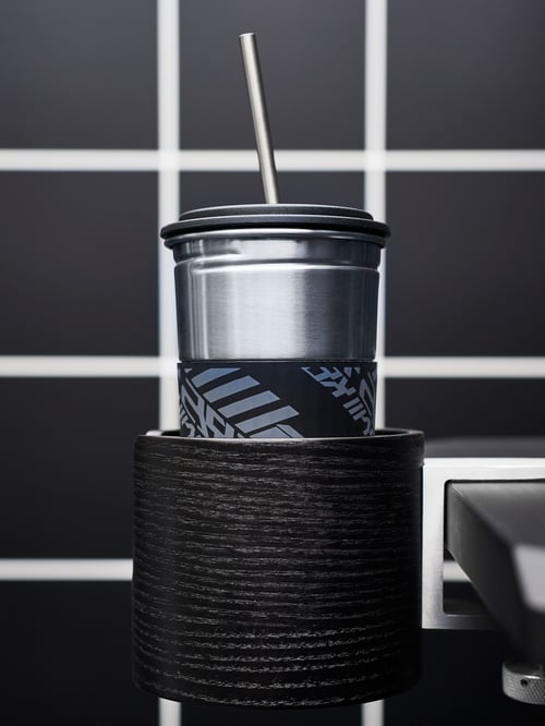 Nahaufnahme eines LÅNESPELARE Becherhalters mit einem LÅNESPELARE Becher mit Deckel und Trinkhalm. Der Becherhalter ist an einer schwarzen Tischplatte befestigt.