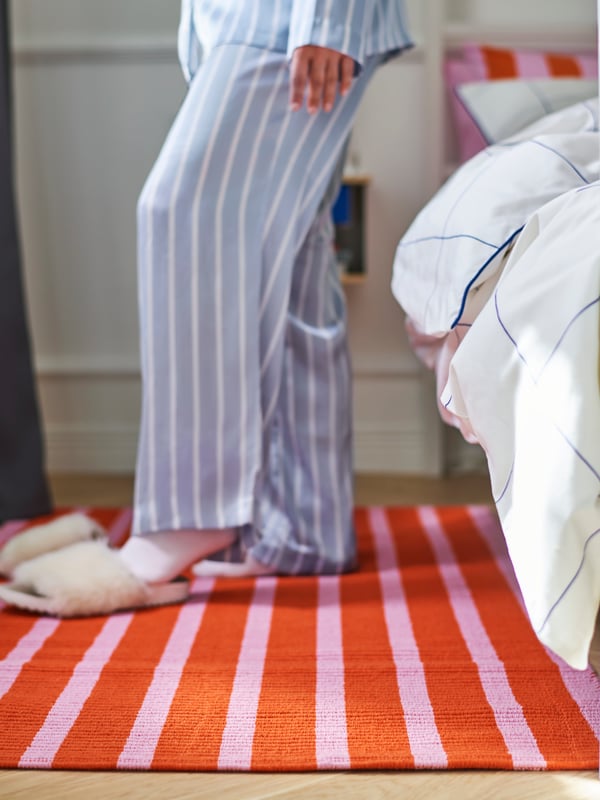 Close-up of VÄGSKYLT rug lying next to a bed, with a person in pajamas standing on it, putting on slippers.