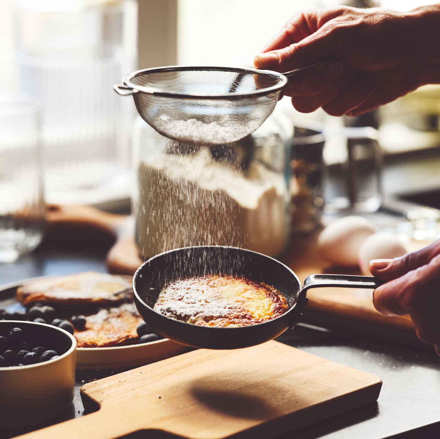 Una persona espolvorea azúcar de un colador sobre un hot cake en un sartén de acero VARDAGEN sobre una tabla para picar PROPPMÄTT.