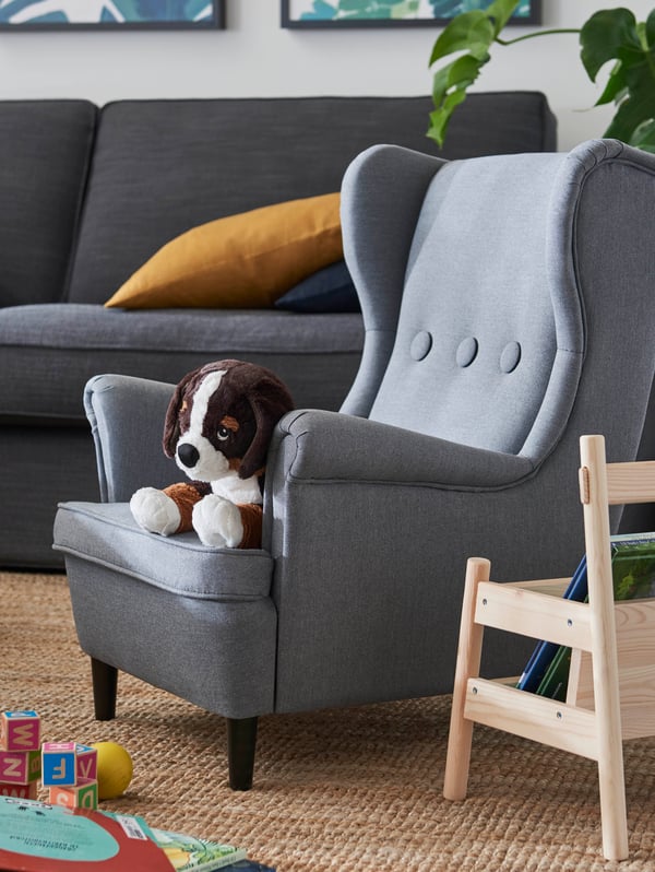 Un sillón infantil STRANDMON color gris oscuro en un living con un sofá grande KIVIK y alfombra LOHALS beige. Sobre el sillón infantil hay un peluche HOPPIG con forma de perro, y se alcanzan a ver otros juguetes a su alrededor. 