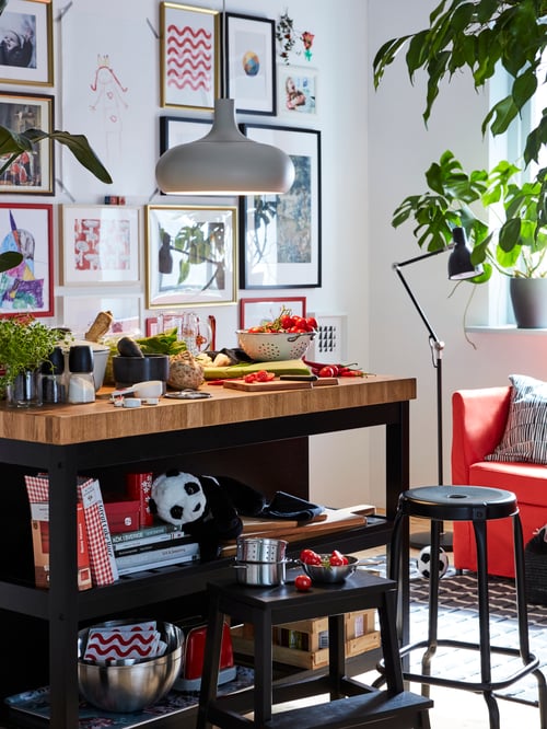A black/oak VADHOLMA kitchen island has books and a soft toy on its shelves. Its butcher block is being used to prep food.