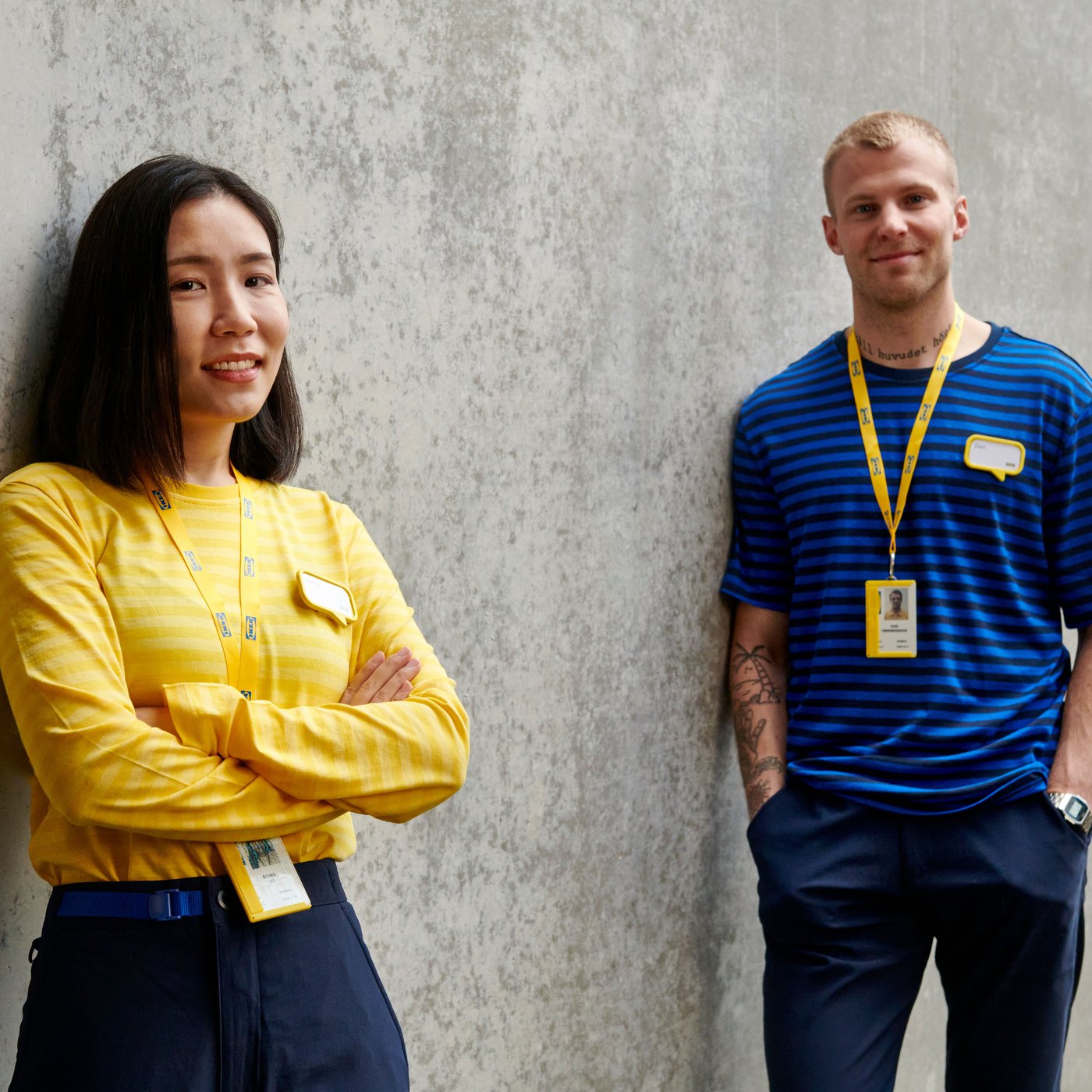 Kaksi IKEA-työntekijää seisovat betonisen seinän edessä, päällään työasut ja katsovat kameraan hymyillen.