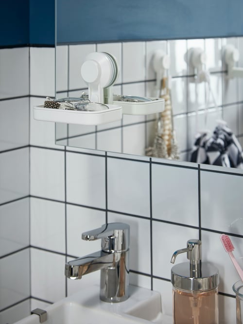 Close-up of bathroom with white tiles, soap dish with suction cup on mirror, liquid dispenser, toothbrush holders, mixer tap.