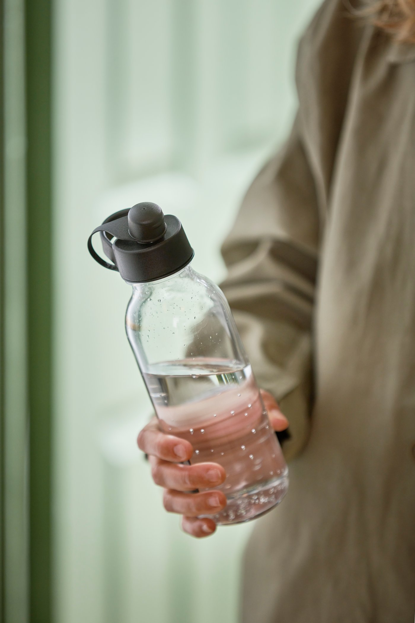 A person in a beige sweater is holding an IKEA 365+ water bottle with a dark grey lid which is half filled with water.