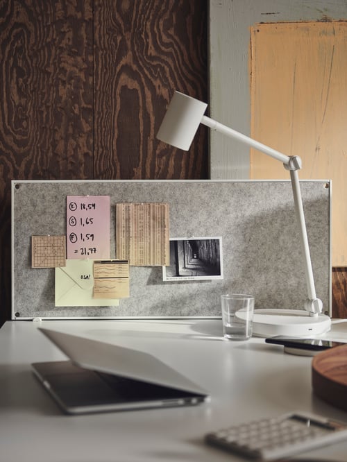 Close-up of workspace with white desk holding laptop, lamp, and grey noticeboard made of steel frame and textile pin board.