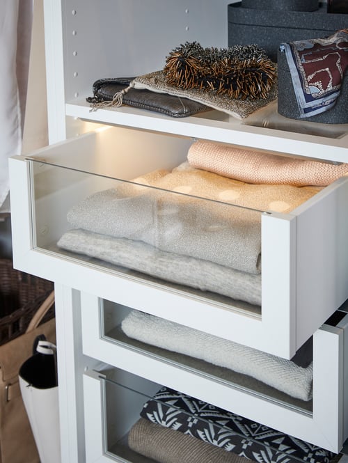 Une armoire-penderie contient des pochettes sur une tablette en verre au-dessus de trois tiroirs KOMPLEMENT blancs avec des façades en verre qui contiennent des chandails. 
