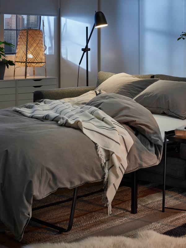 VIMLE sofa-bed unfolded to a bed, with mattress, quilt and pillows, with evening city view in the background.