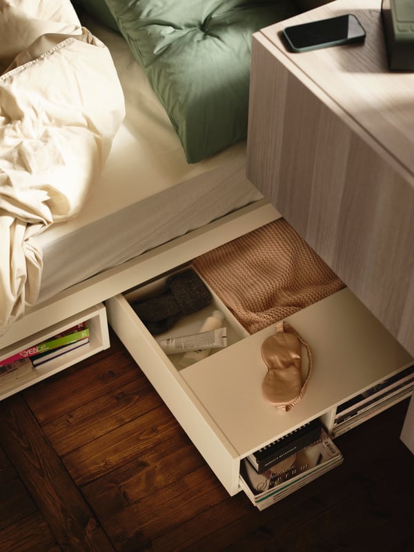 A white FREDVANG underbed storage/bedside table wheeled out from below a bed dressed in ÄNGSLILJA bed linen.