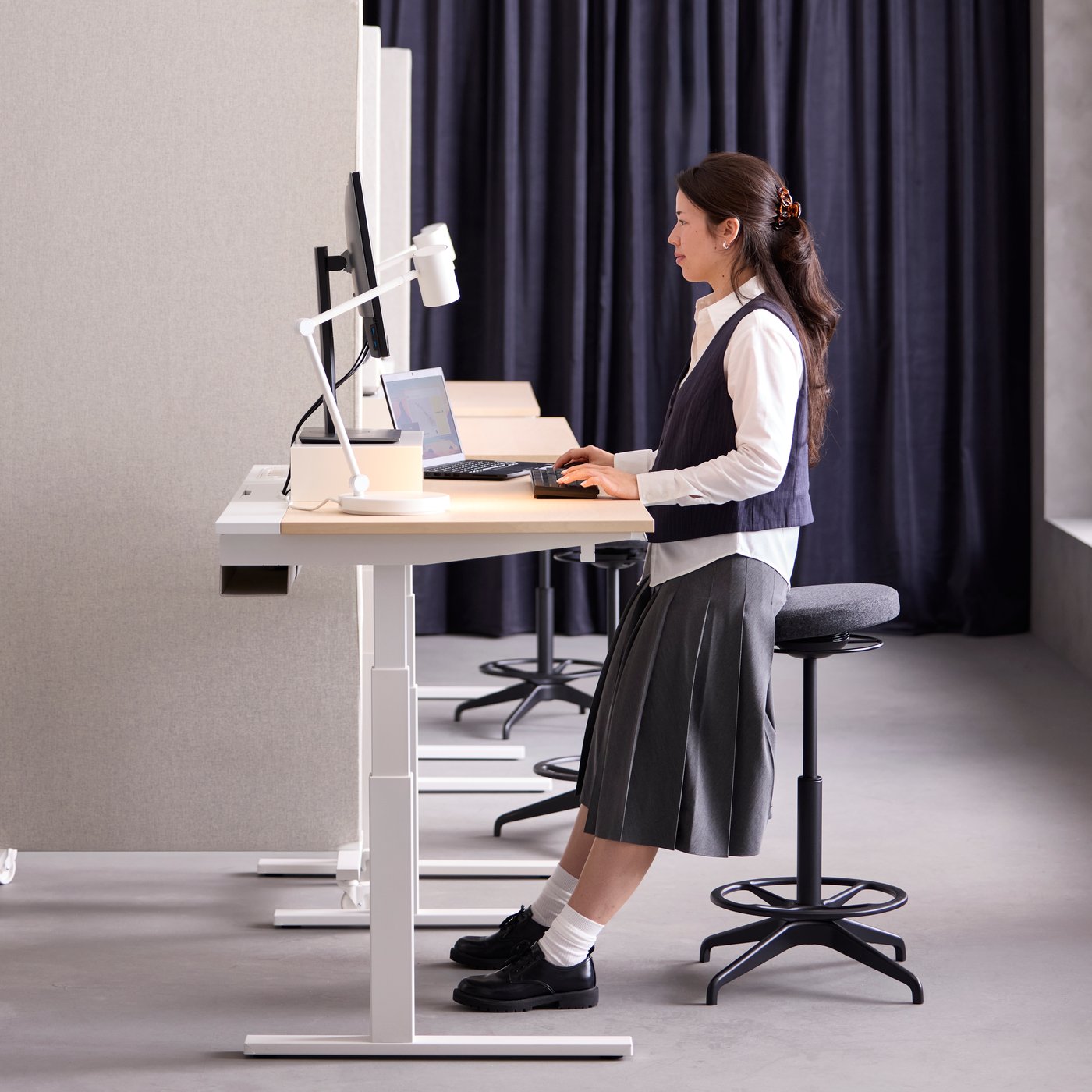 White MITTZON birch veneer sit/stand desk in mid-high mode with a person leaning on sit/stand support while working.