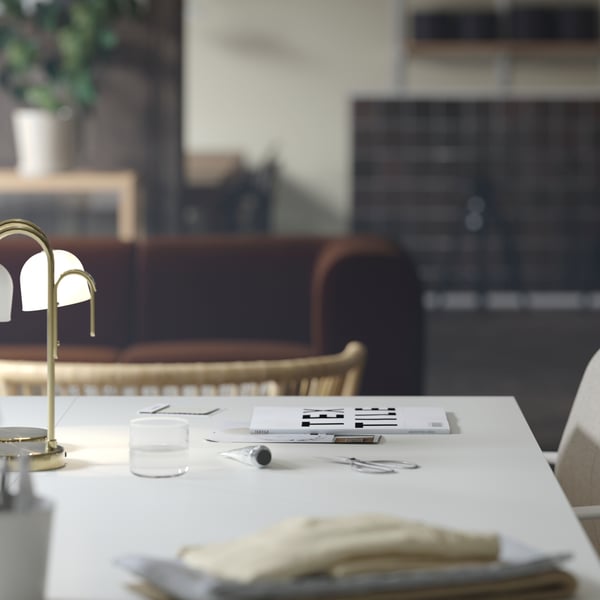 A close-up of a white TROTTEN desk with a TVÅMASTAD LED portable lamp on top. A dark brown sofa is visible in the background.