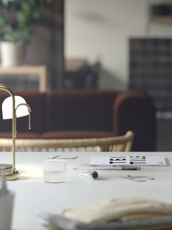 A close-up of a white TROTTEN desk with a TVÅMASTAD LED portable lamp on top. A dark brown sofa is visible in the background.