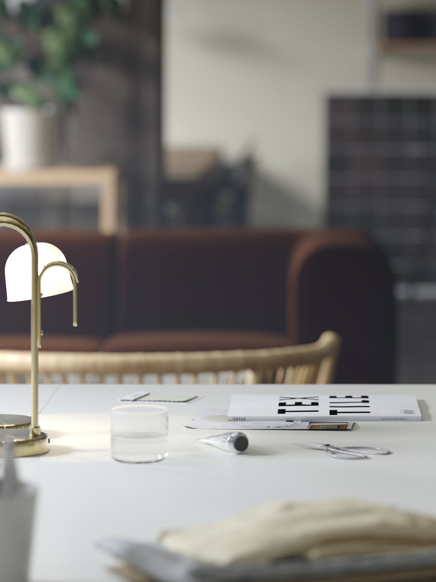 Es ist eine Nahaufnahme eines weissen TROTTEN Schreibtisches mit einer tragbaren TVÅMASTAD LED-Leuchte darauf zu sehen. Dahinter steht ein dunkelbraunes Sofa.