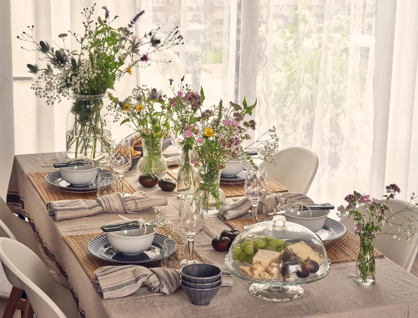 Festlich gedeckter Tisch mit Geschirr, Gläsern und mehreren Vasen voller Wildblumen in hellem Raum.