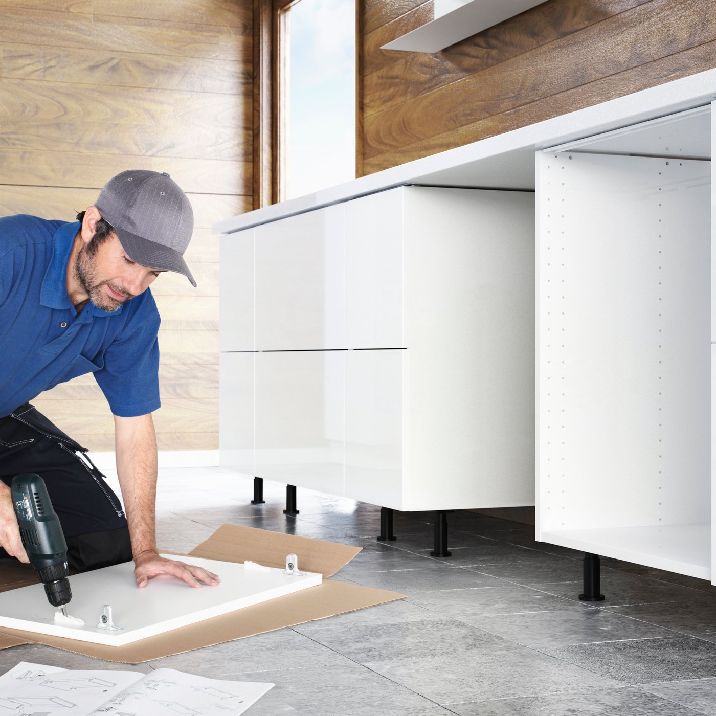 Kitchen installation service: A person in working gear kneeling on a tiled floor in a partially finished kitchen, drilling screws into a white front.