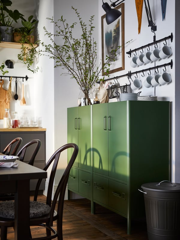 Two dark green IDÅSEN cabinets with doors and drawers are against a dining area wall. Above them, many mugs hang from rails.
