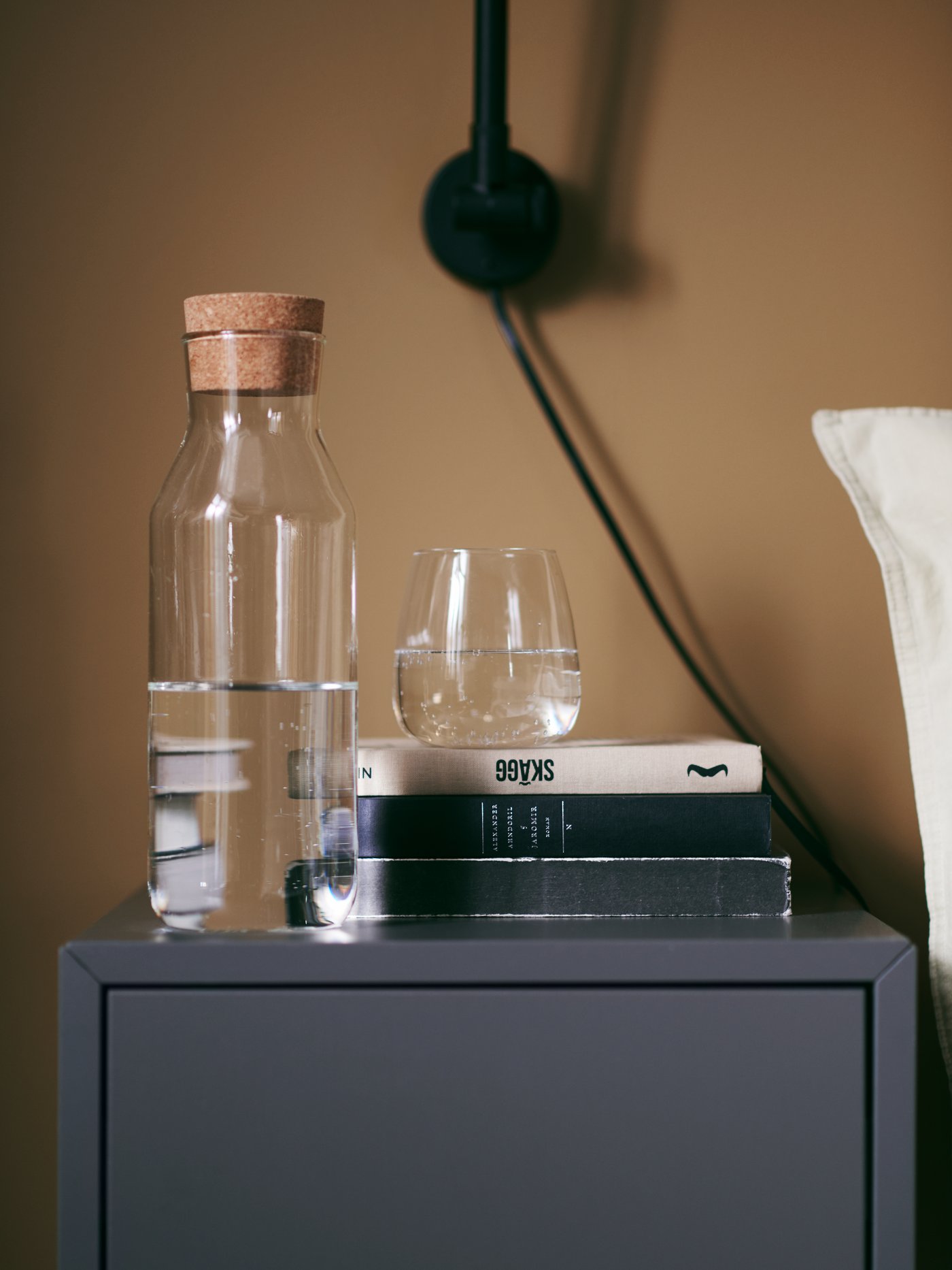 Una garrafa llena de agua con tapa de corcho y un vaso sobre una mesita de noche negra.