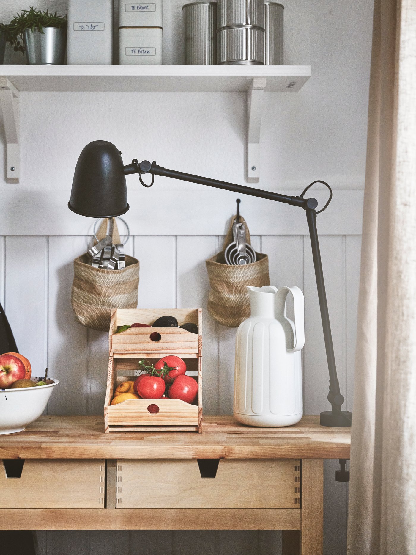 Träbord med en trälåda fylld med frukt framför en bordslampa med lång, justerbar arm. På bordet står även en vit kanna. Ovanför bordet finns en hylla med burkar och två hängande korgar.