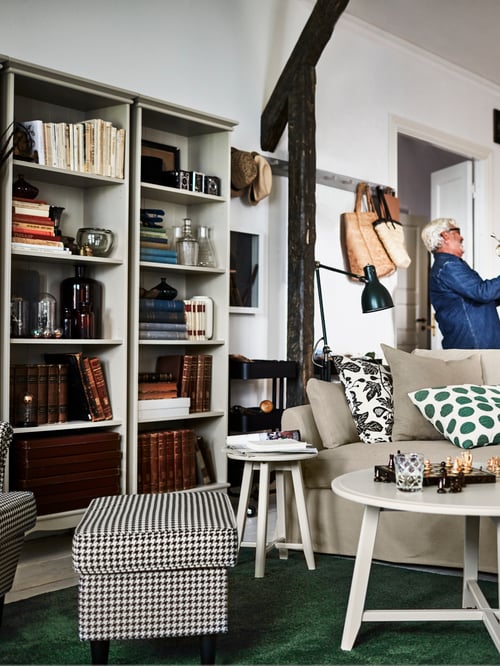 Wohnzimmer mit grünem Teppich, Hocker, Bücherregal, Arbeitsleuchte, zwei Satztischen und einem Sofa mit mehreren Kissen.