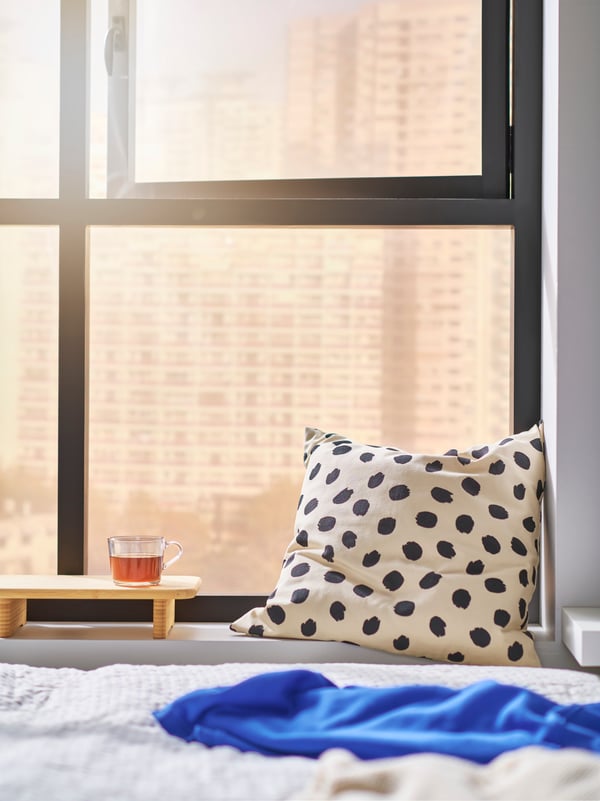 Vista parziale di un letto in primo piano, una grande finestra sullo sfondo e, sul davanzale, un cuscino ODDNY a pois bianchi e neri e un vassoio.