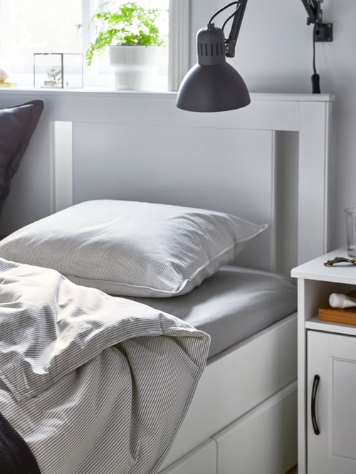 A grey wall lamp is above a partially visible, white SONGESAND bed frame with four storage boxes and grey, striped bedding.