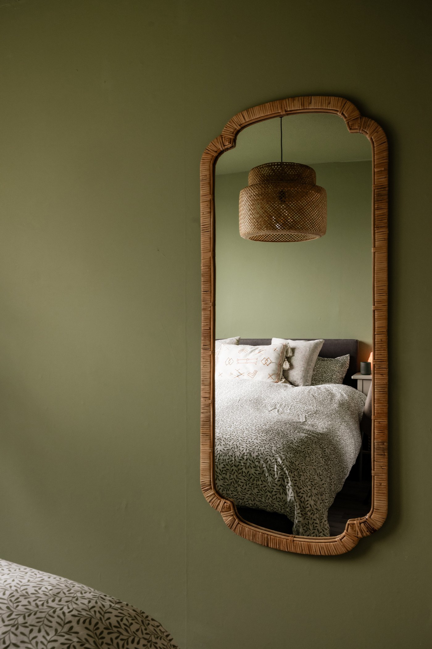 Een zachtgroene muur in de slaapkamer waar een rotan KANDLENÖT spiegel hangt. In de spiegel zie je het bed en de hanglamp.