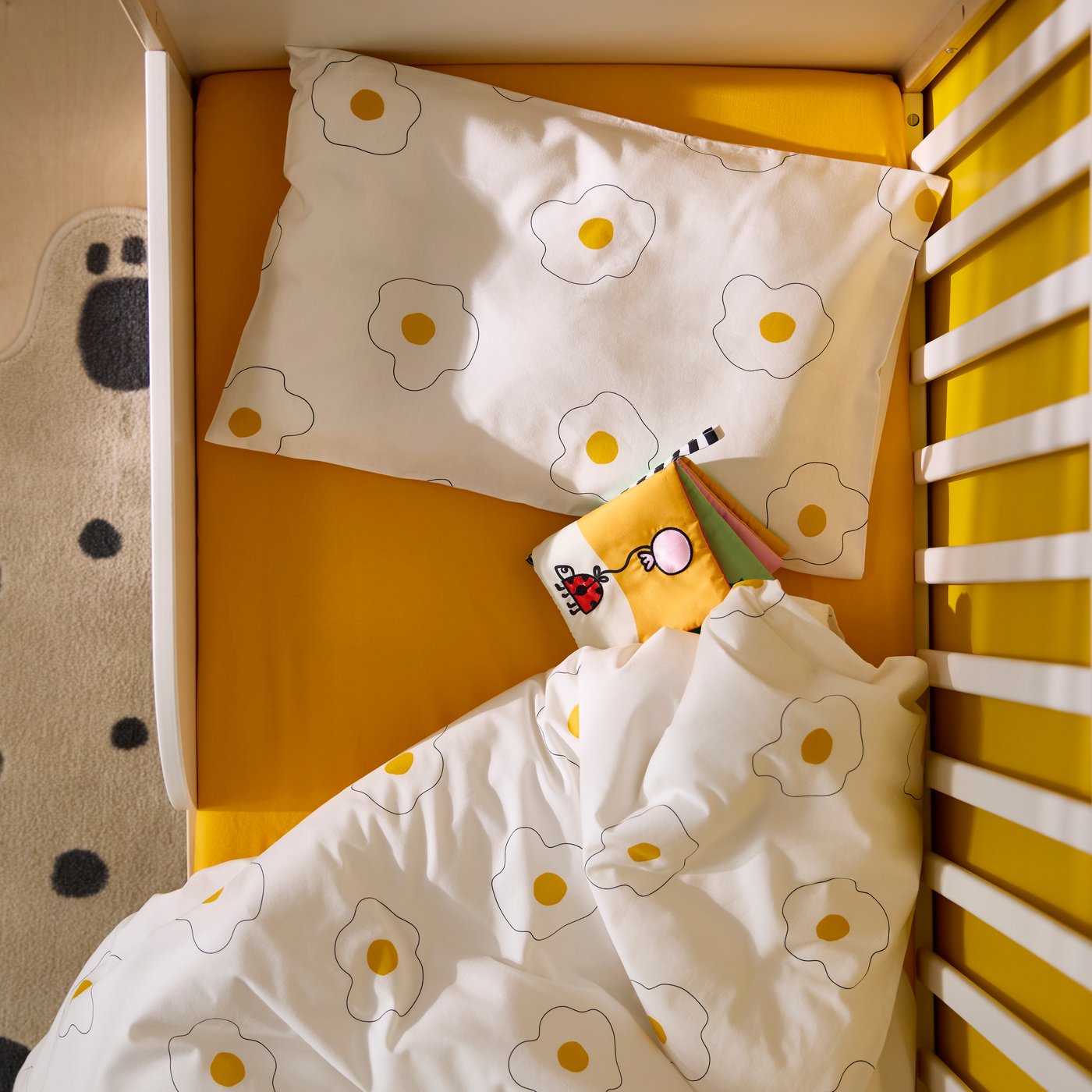 Vue en plongée sur un lit d’enfant avec un drap jaune et une housse blanche à motifs d’œufs au plat.