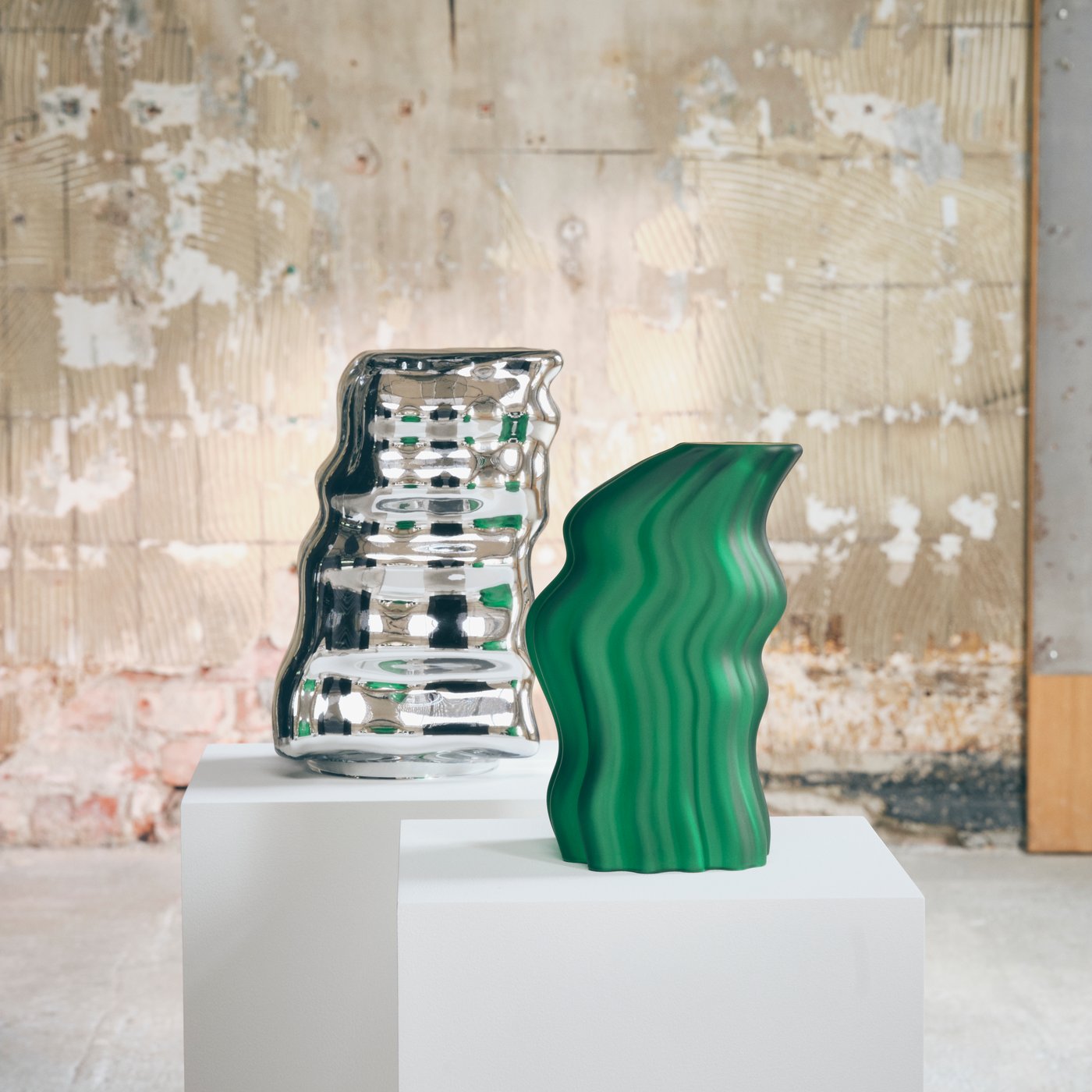 Close up of a green and a metallic art object standing on white pedestals agaist a rough concrete wall.