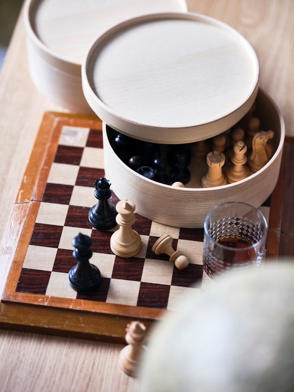 A MALLGRODA round storage box filled with chess pieces, standing on a chessboard.