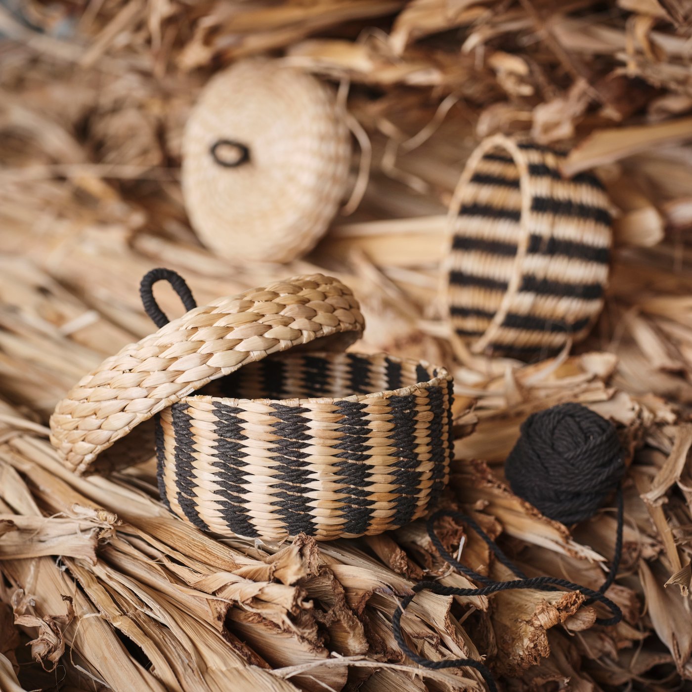 A black/beige striped MÄVINN box with lid sits on a pile of banana bark fibres. Next to it lies a ball of black cotton.