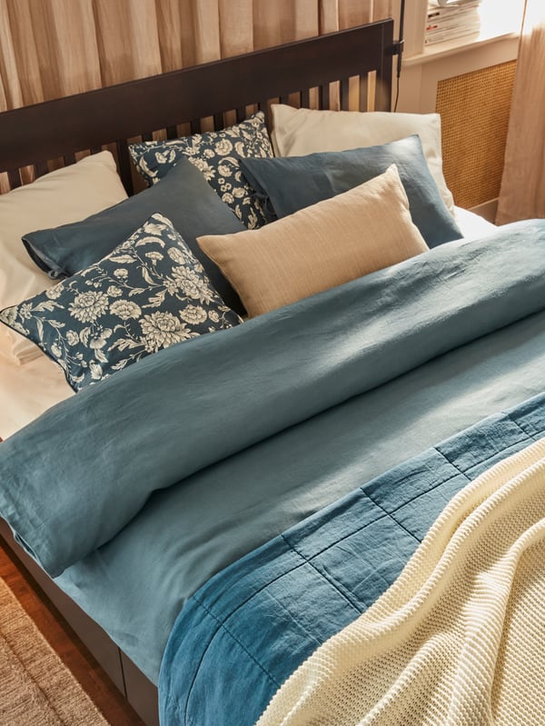 A bed with plenty of pillows in blue, beige and white, a dark blue quilt cover, off-white knitted throw, and linen curtains.