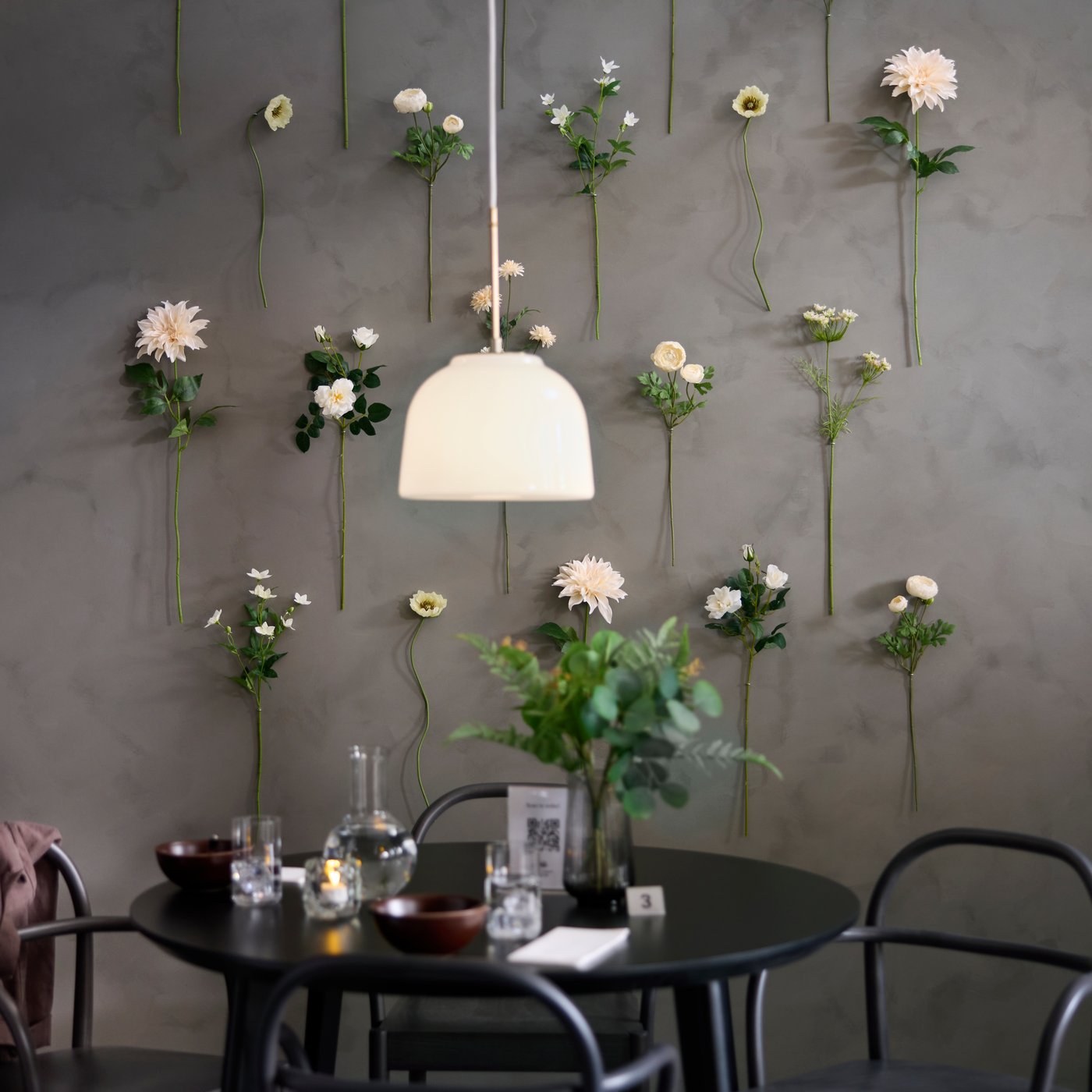 Jídelní kout v kavárně s aranžmá z umělých květin SMYCKA zavěšených na stěně nad černým kulatým stolem LISABO.