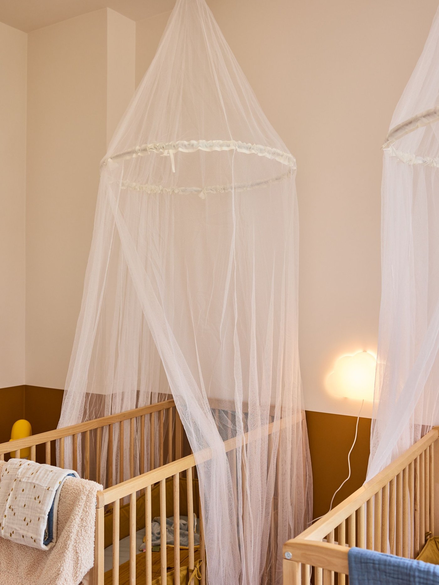 Twee houten babybedjes met witte klamboe, zacht beddengoed en warme verlichting in een rustige, gezellige babykamer.