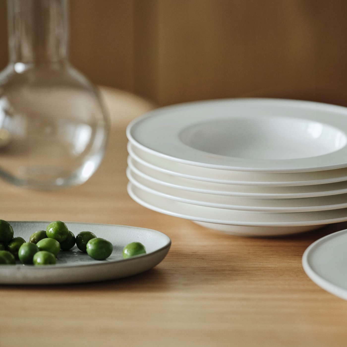 A tabletop with a grey STOCKHOLM 2025 serving plate with food next to a carafe and five white stacked deep plates.