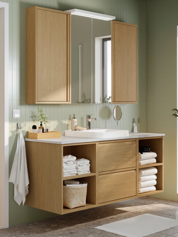 A pale green bathroom has an ÄNGSJÖN vanity sink in oak effect with drawers and open shelves and a mirror cabinet above it.