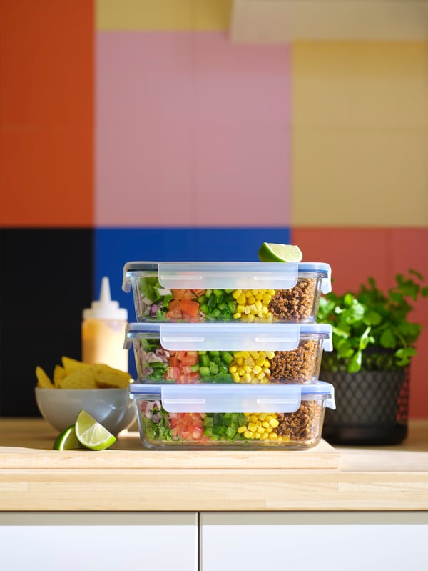 Drei gestapelte IKEA 365+ Lebensmittel Behälter mit Salatzutaten auf einer Arbeitsplatte vor einer bunten Wand.
