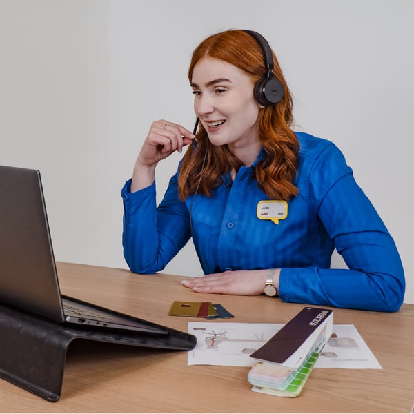 Projektantka IKEA siedząca przed laptopem przy biurku w niebieskiej koszulce z zestawem słuchawkowym na głowie. Prowadzi zdalnie usługę indywidualnego projektowania wnętrza dla firm i trzyma palcami mikrofon. Na blacie stołu leży wzornik kolorów i wydruk kolażu produktowego.
