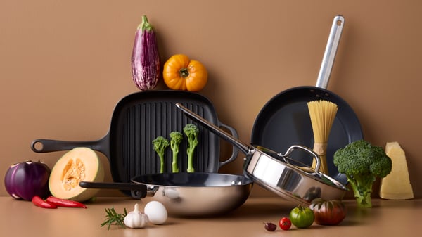 Four pans of different styles and materials are placed on a brown surface with different kinds of vegetables around them.