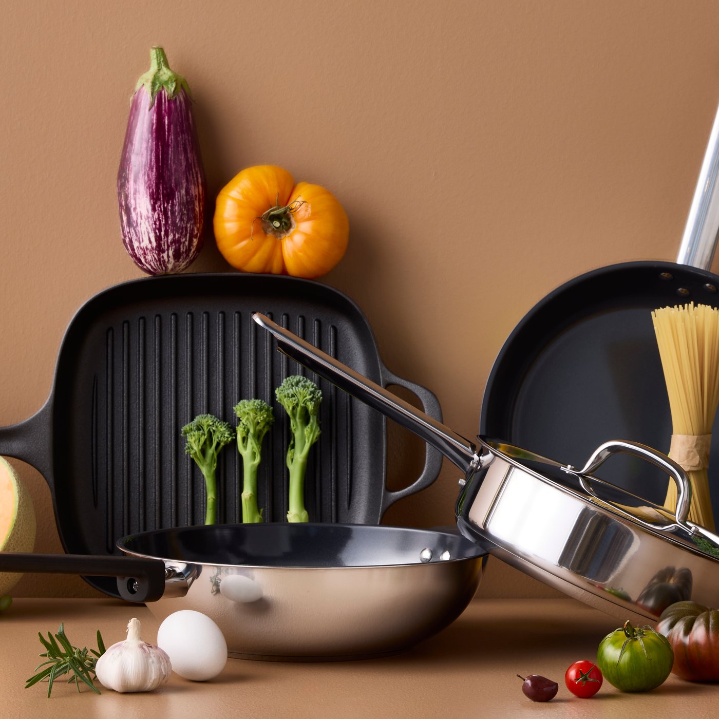 Four pans of different styles and materials are placed on a brown surface with different kinds of vegetables around them.