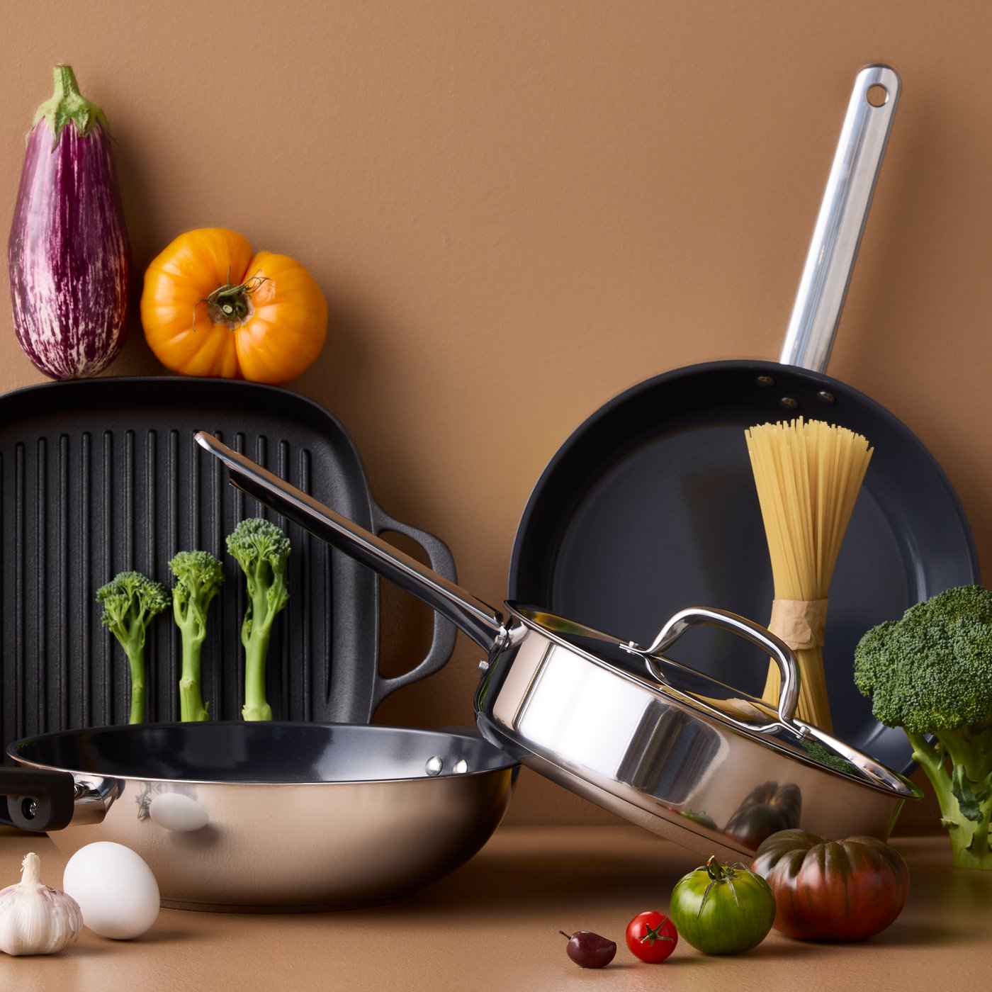 Four pans of different styles and materials are placed on a brown surface with different kinds of vegetables around them.