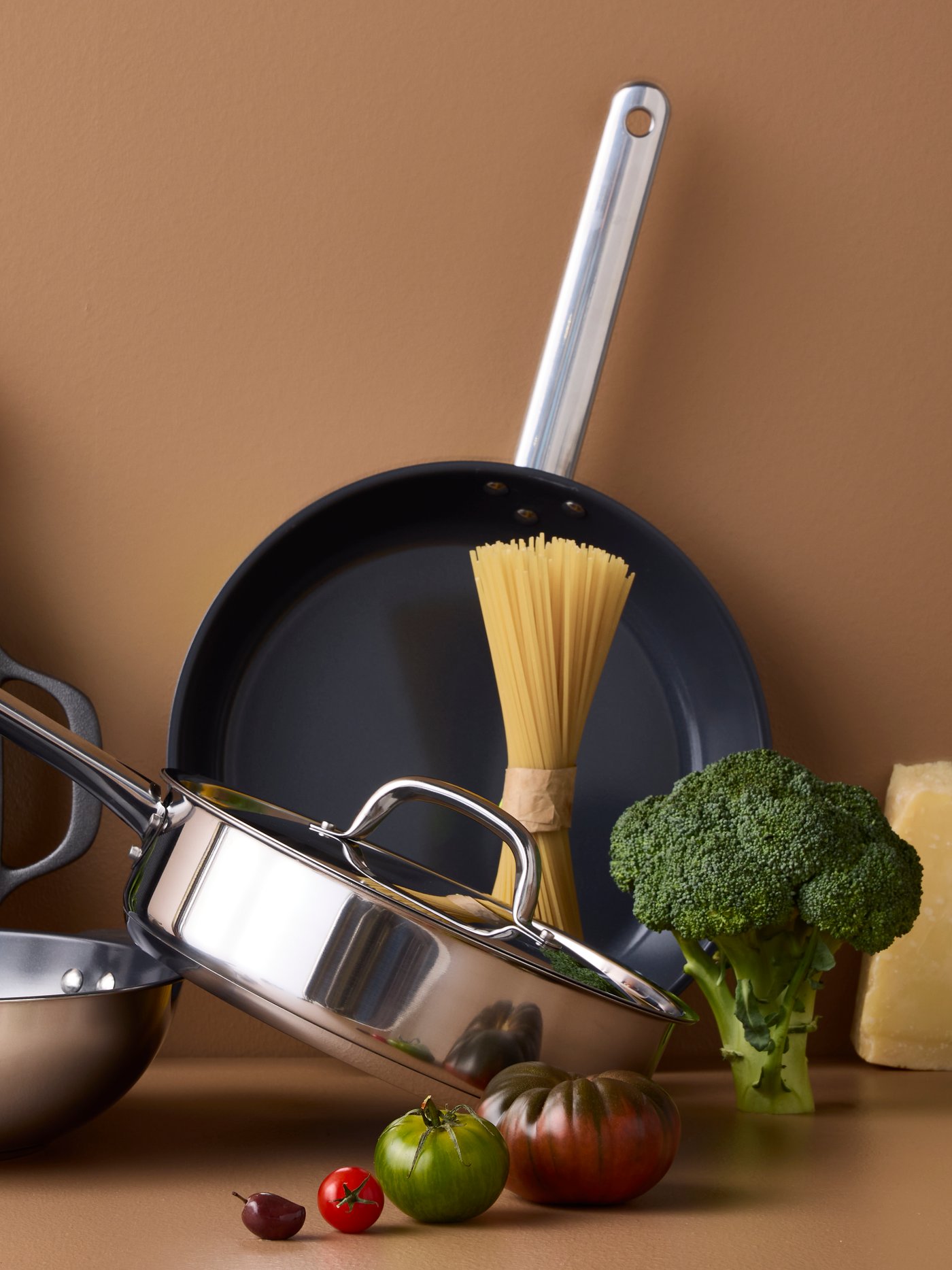 Four pans of different styles and materials are placed on a brown surface with different kinds of vegetables around them.