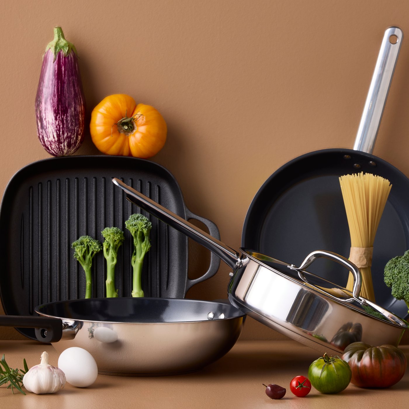 スタイルや素材の異なるフライパン4個がブラウンのテーブルの上に置いてある。まわりにさまざまな種類の野菜。