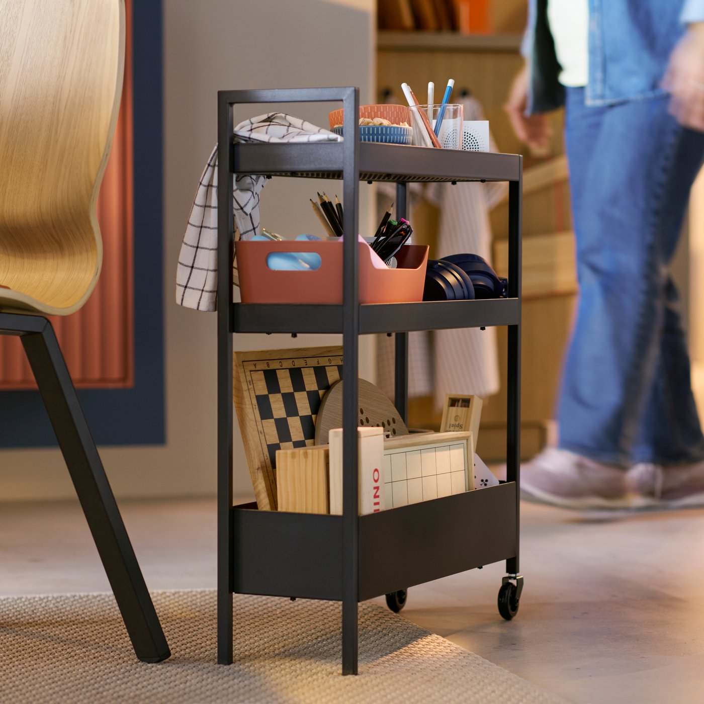 A black NISSAFORS trolley has items stored on it, including some games, and a person in the background is walking past it.