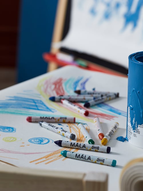MÅLA mixed-colours wax crayons scattered across a sheet of paper with colourful drawings on it. A MÅLA easel stands nearby.
