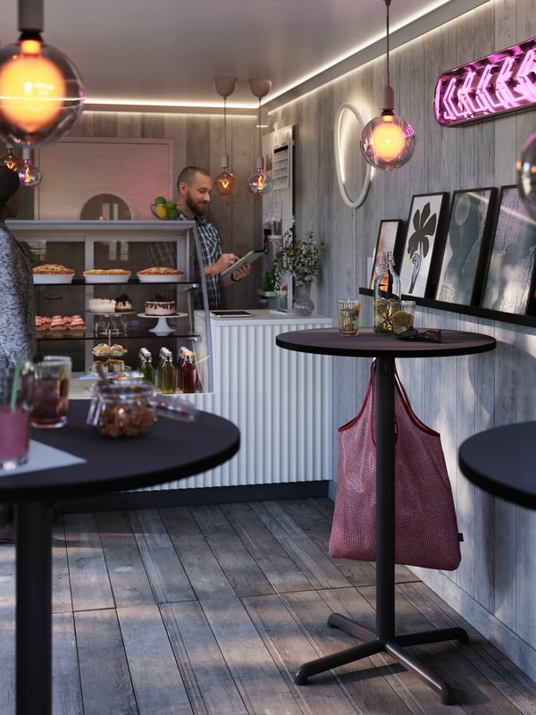 A person is standing behind the counter at a café with STENSELE bar tables, next to a display case, holding a mobile device.