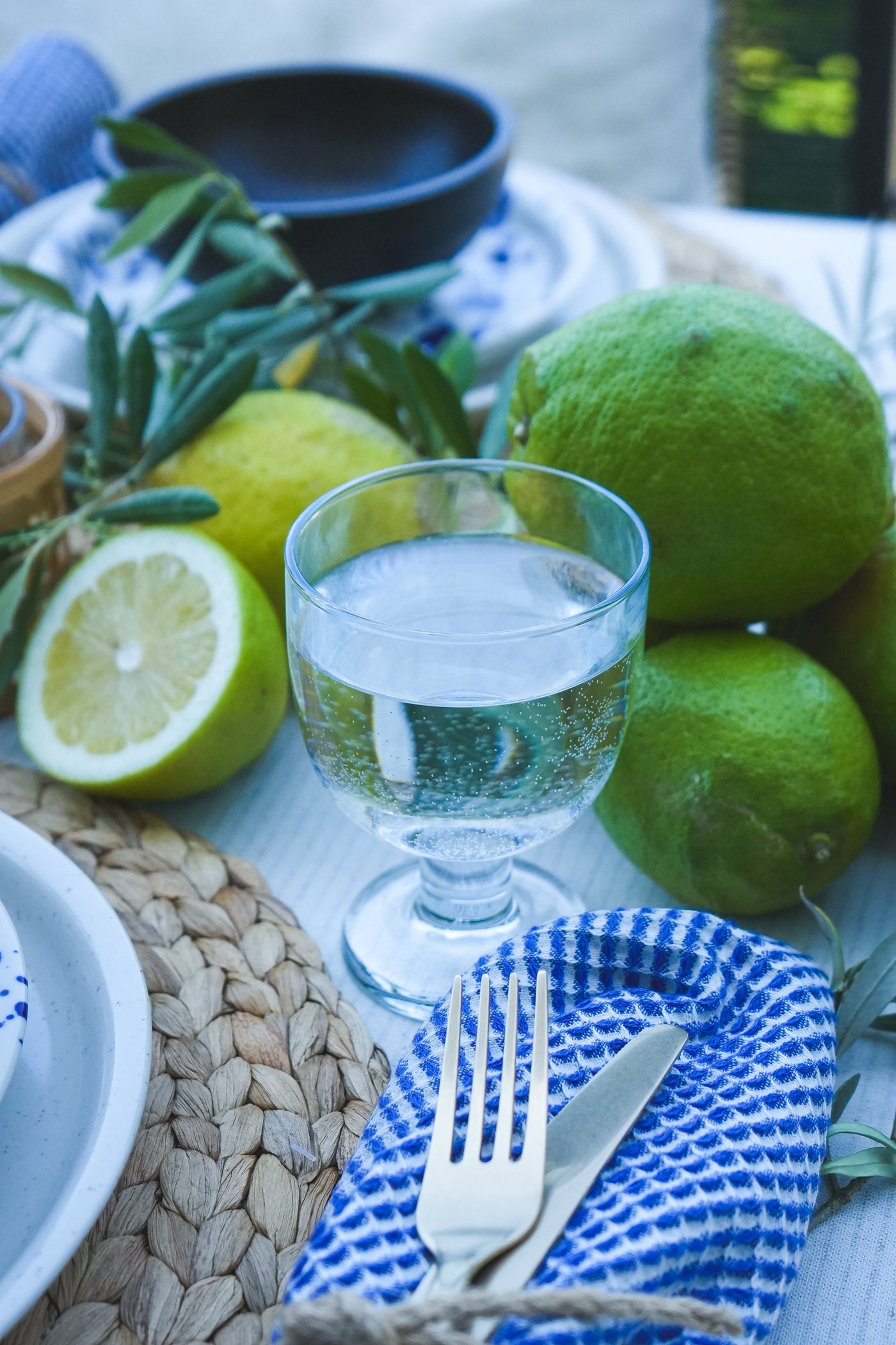 Une table dressée avec un verre d'eau FRUKTDUVA, des couverts enveloppés dans un linge bleu et blanc, des citrons verts frais et des brins d'olivier en guise de décoration méditerranéenne.