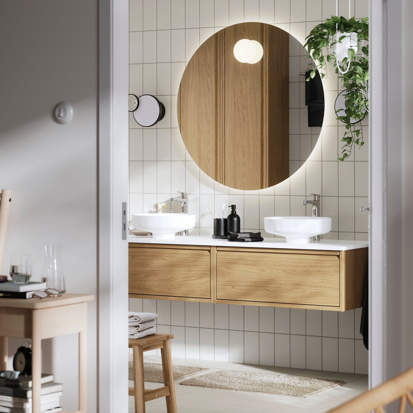 Łazienka z okrągłym, podświetlanym lustrem, dwoma umywalkami, drewnianą szafką pod umywalki i ścianą wyłożoną płytkami. Zieleni dodaje wisząca roślina.