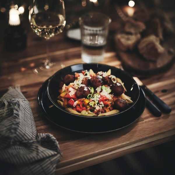 A pasta and meatballs dish in a GLADELIG bowl on a table surrounded by a napkin, a water glass and a glass of sparkling drink.