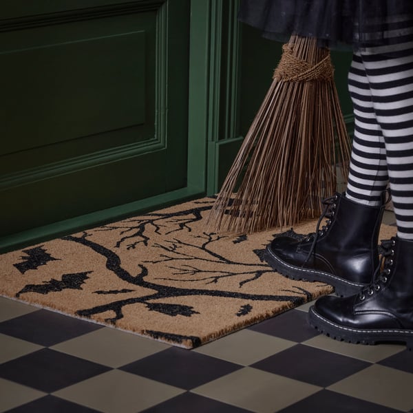 A kid wearing boots and striped tights steps on a HÖSTAGILLE door mat with a bat design by a green door.