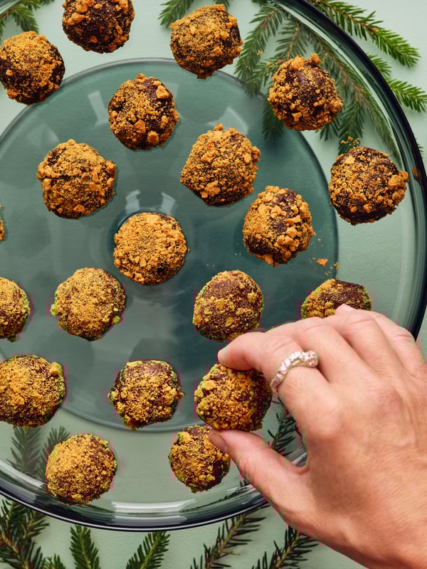 Una mano está cogiendo un dulce de una bandeja de cristal llena de trufas doradas y cubiertas de migas, colocada sobre una superficie verde.